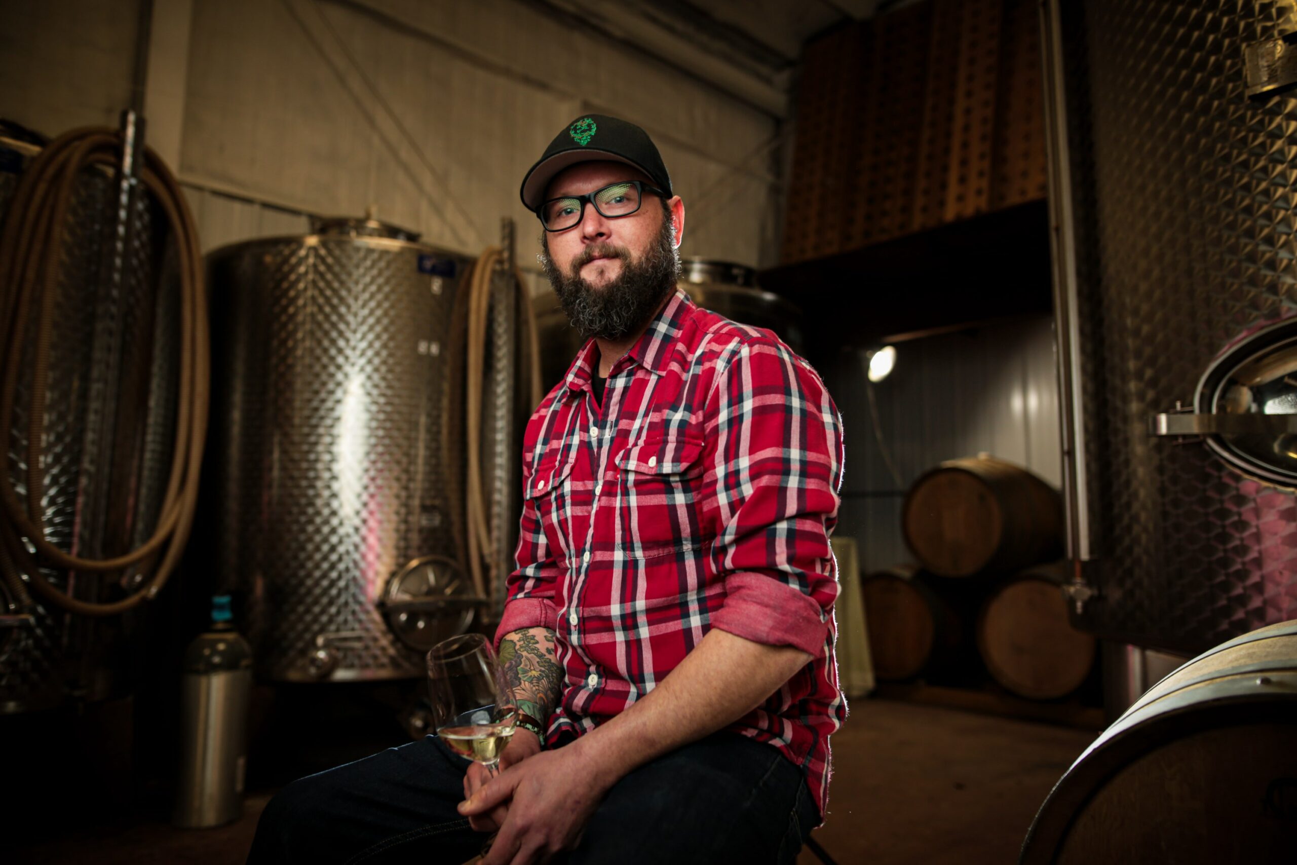 Winemaker Nathan Kendall in front of tanks. Man in red flannel shirt with black baseball cap and glasses, holding wine glass.