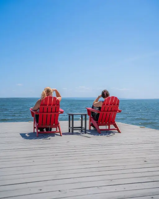 Get RED-y Vacations two women sitting in red Adirondack chairs lakeside
