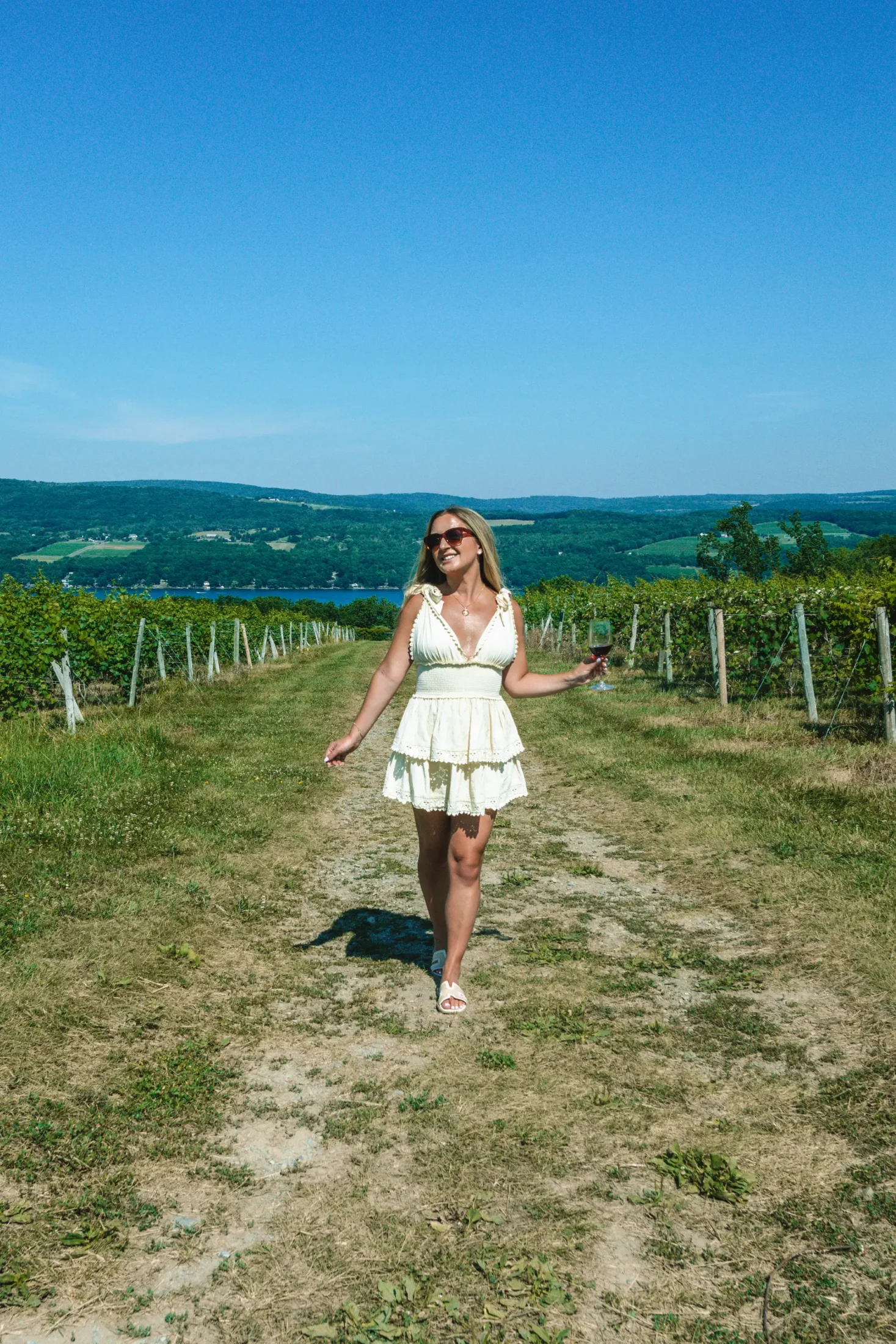 Woman walking through a vineyard with wine in her hand and Seneca Lake in the background.